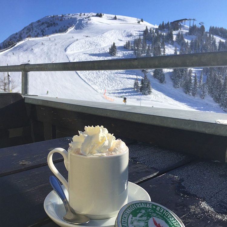 En kopp med varm choklad står på ett bord som blickar ut över en skidbacke i alperna.